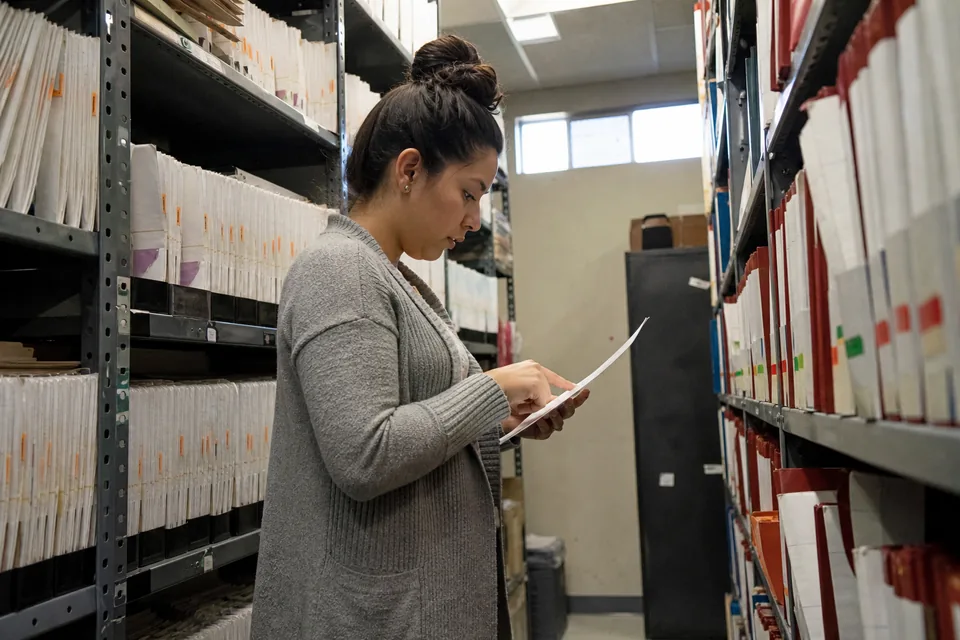Paralegal standing between shelves of labeled case binders in a law firm records room, checking a date on a sheet of paper