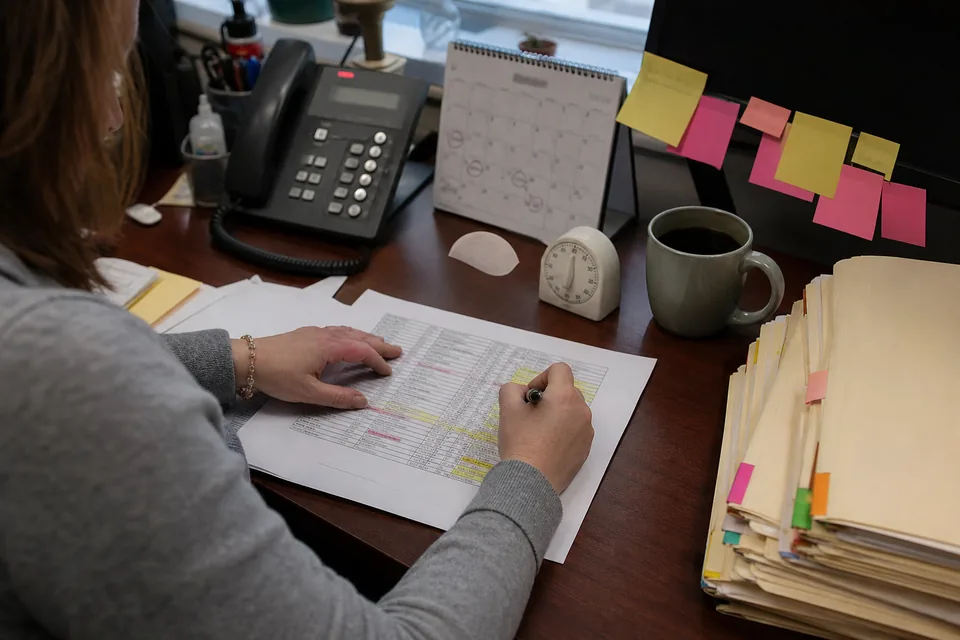 Firm administrator's desk crowded with a spreadsheet printout, a kitchen timer, sticky notes, a paper calendar and a stack of client folders